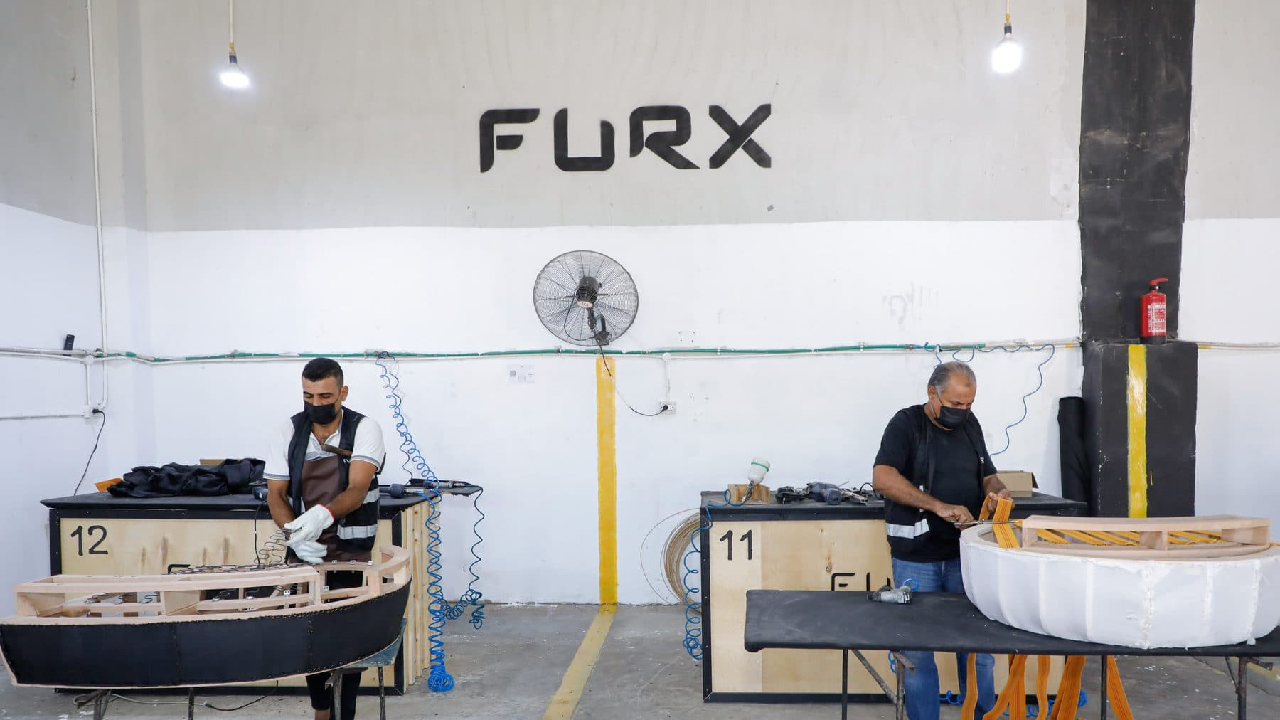 Two masked upholsterers at branded FURX stations fit springs and trim orange elastic webbing on curved sofa frames.