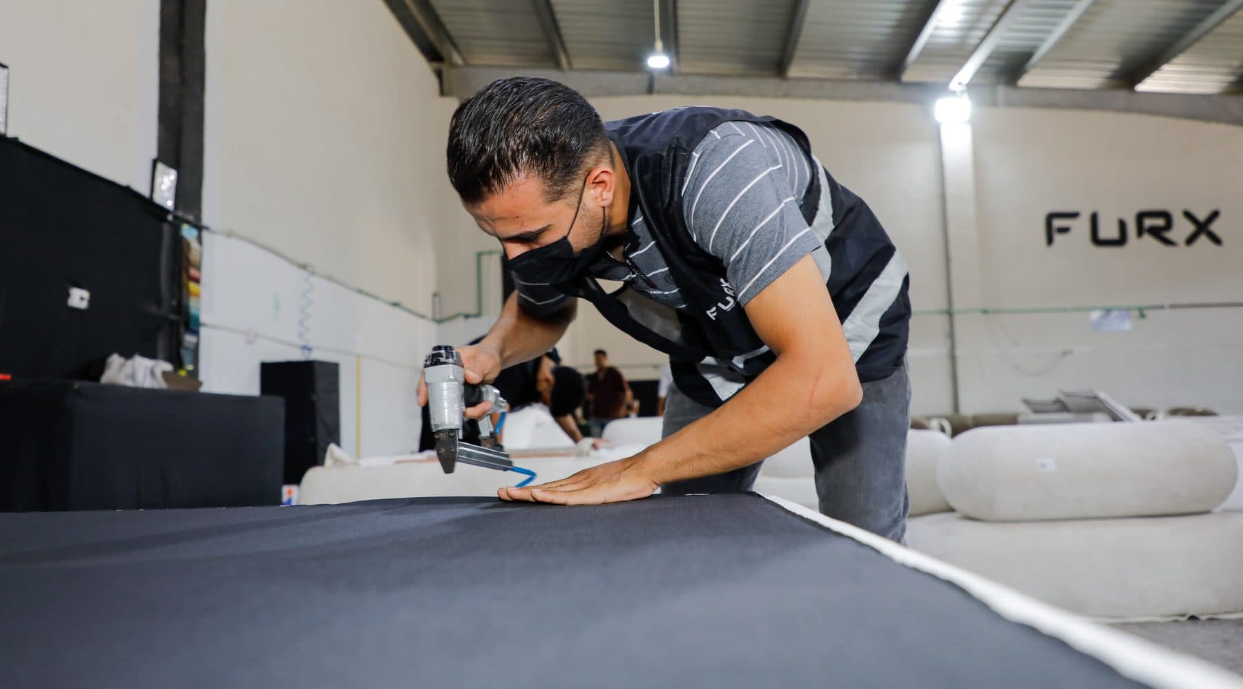 An upholsterer staples black dust-cover cloth to the underside of a sofa body with a pneumatic stapler.