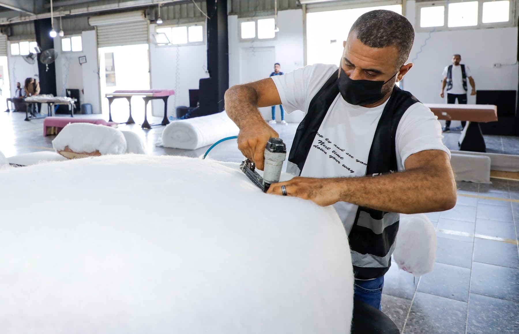 An upholsterer in a mask staples polyester wadding over a rounded sofa form with a pneumatic stapler.