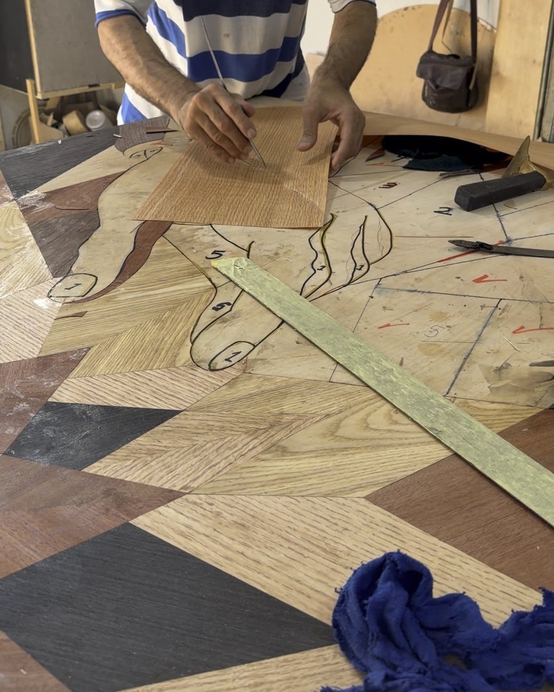 A craftsman holds a leaf of light veneer over the drawn outline of a hand on the marquetry base, with laid pieces and a steel straightedge around it.