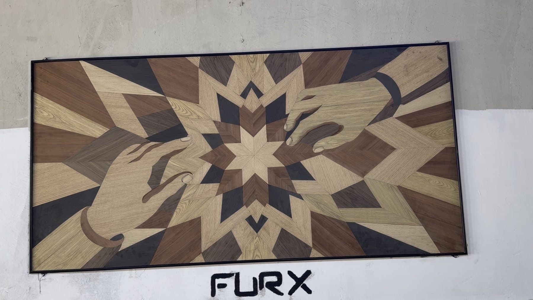 A framed wood-veneer marquetry panel on a factory wall: a star in light and dark woods at the centre, with two hands reaching in from either side, above the FURX letters.